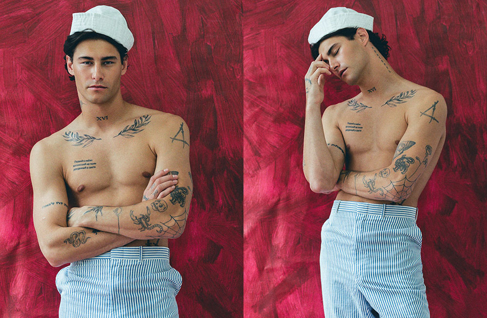 Tattooed shirtless man in a sailor hat and striped pants against a red backdrop.